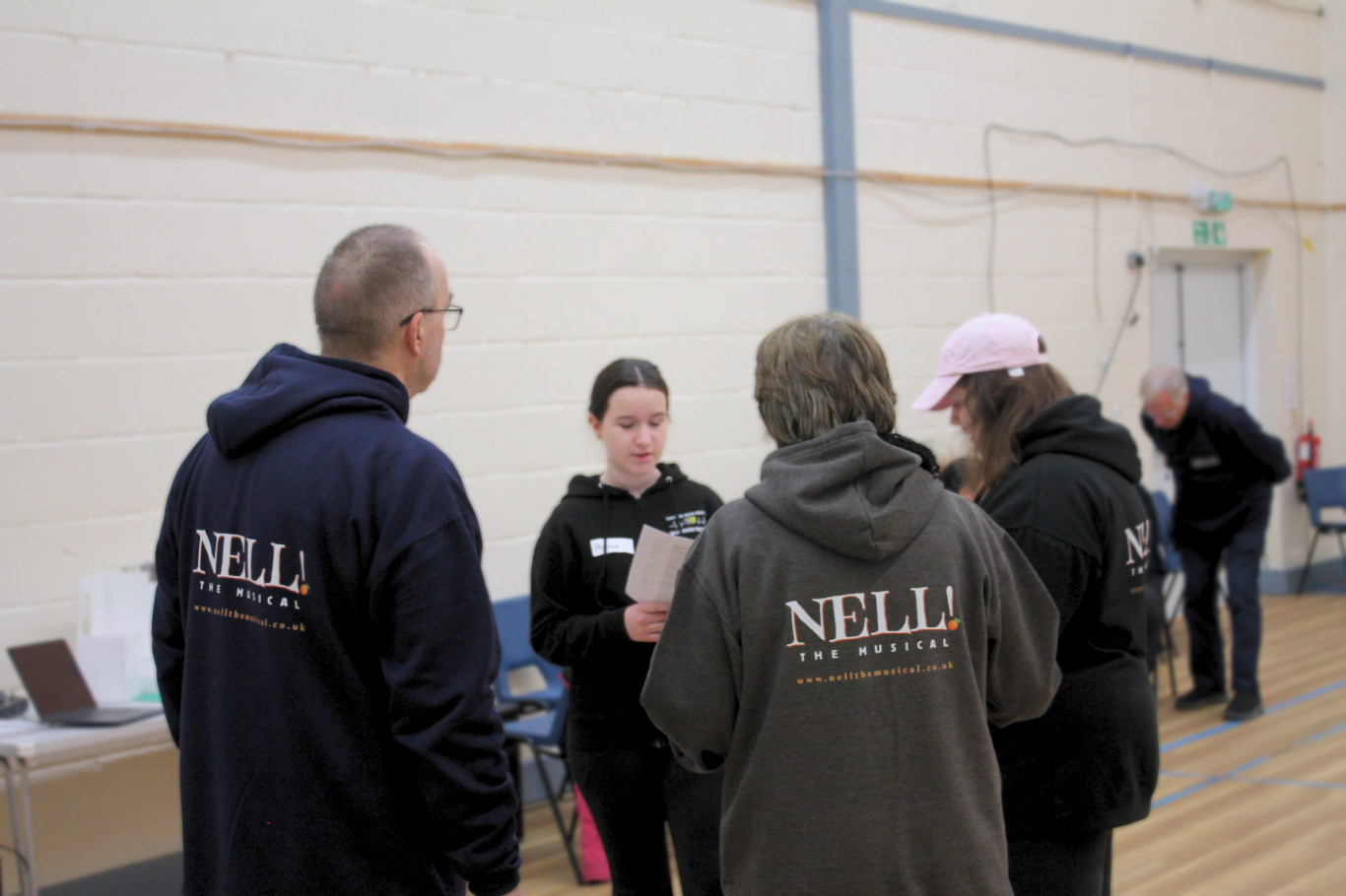 A pre-teen girl reads from a piece of paper. Three adults are huddled around her wearing Nell! The Musical hoodies.