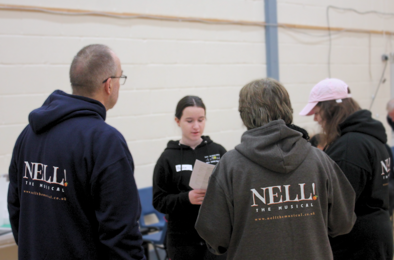 A young girl reading from a sheet of paper with adults wearing Nell! The Musical hoodies huddled around her.