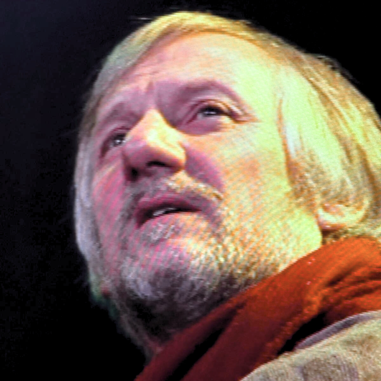 Close up of man in a theatre setting with light coloured hair and beard wearing a red scarf looks to the distance.