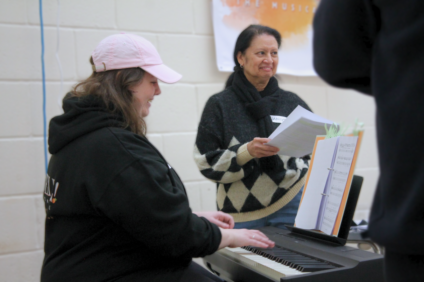 Two smiling women, one plays an electric keyboard, the other stands beside holding sheets of paper.