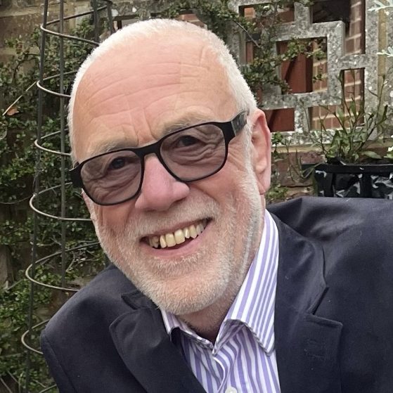 A man in an outdoor setting with very short white hair and beard, wearing dark glasses, smiles at the camera.
