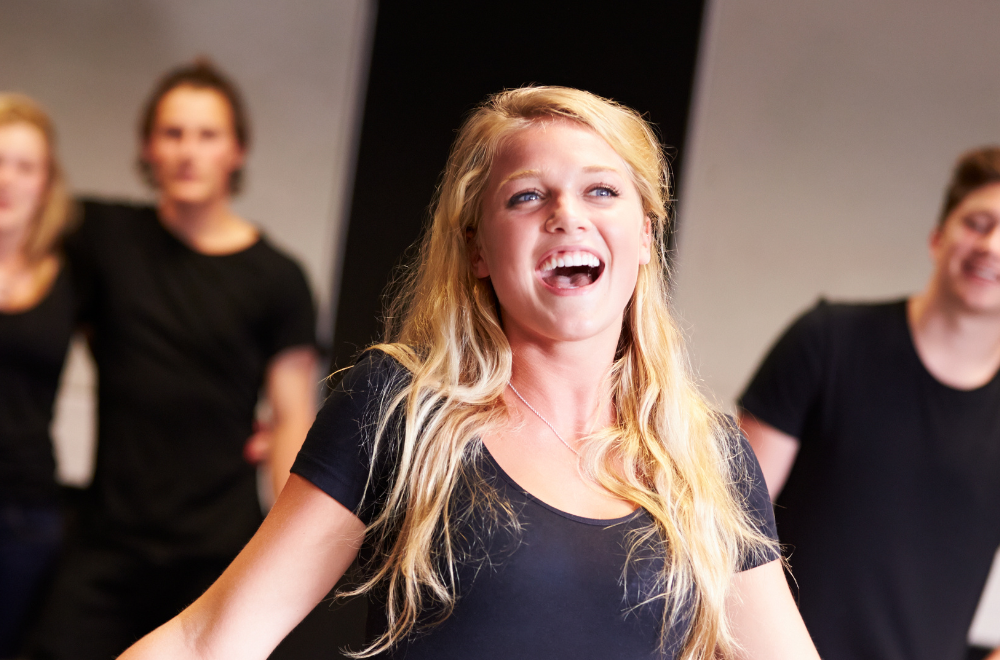 A young woman with long blonde hair singing in the front of a small group of people wearing black.
