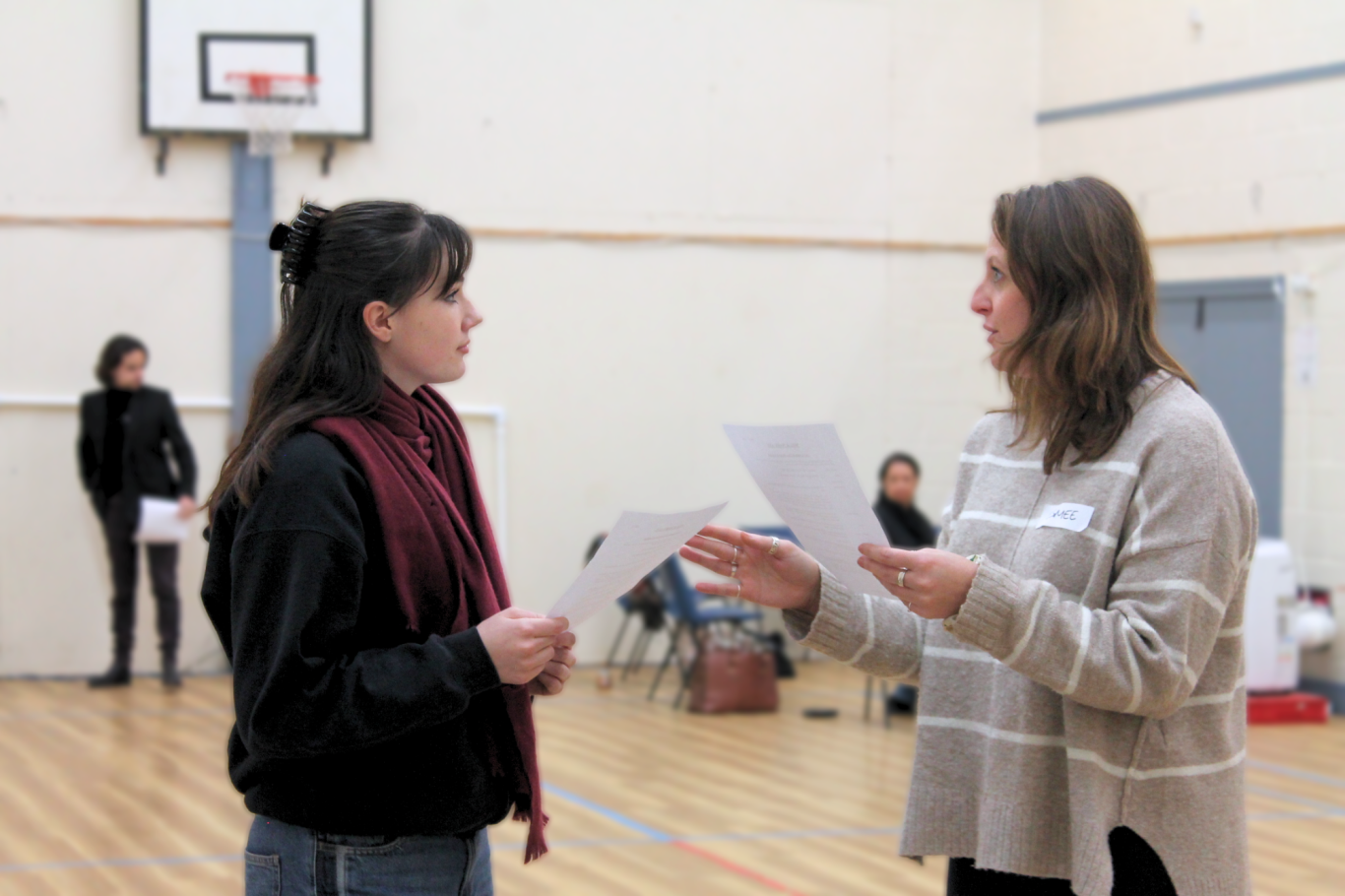 Two young women stand facing each other in a gymnasium. They are conversing while holding pieces of paper.