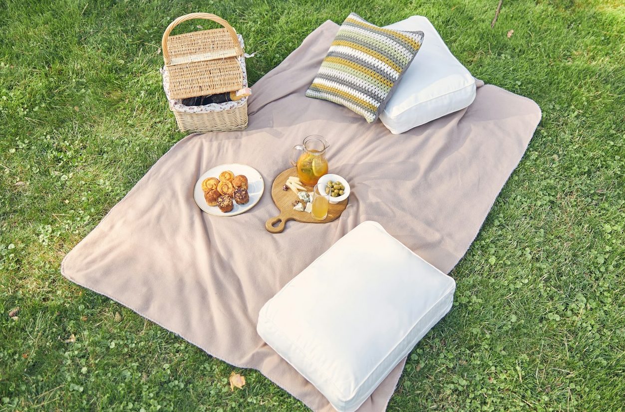 A beige picnic blanket on green grass with cushions, a picnic hamper and two plates of food.
