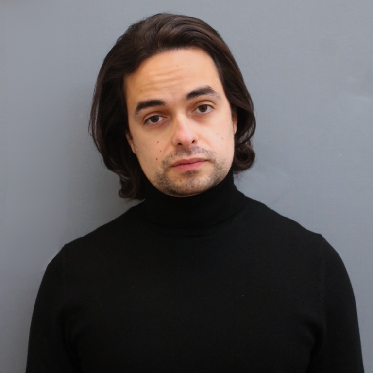 A young man with medium length dark hair and stubble looks towards the camera.
