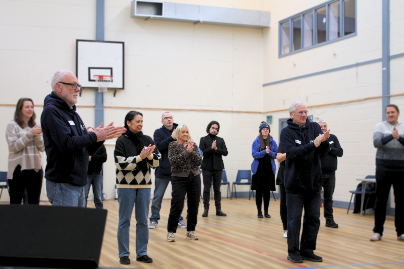 A group of people standing in a gymnasium, most are smiling. They are rubbing their hands together.