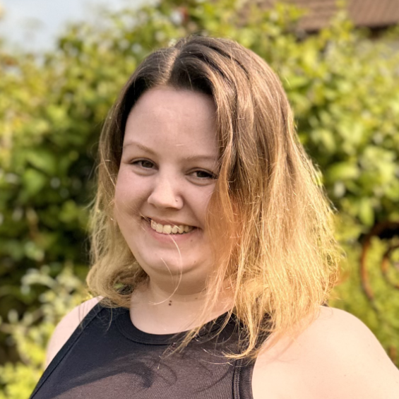 A young woman on a background of greenery looks to the camera with a laughing expression.