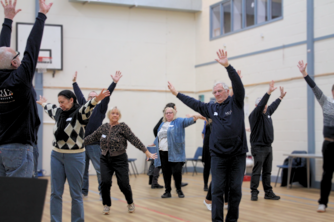 Group of people in a gymnasium setting reaching their arms up in the air.